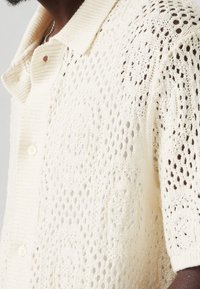 Man met een crèmekleurige, kortslijf haakgebreide cardigan met knoopsluiting aan de voorkant en getextureerde cirkelvormige patronen.