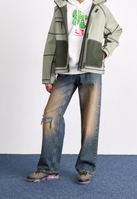 Person som bär en grön och grå jacka, vit hoodie med gröna löv och röd logotyp, blekta vida jeans med knäpatch, och gröna sneakers med lila sulor.