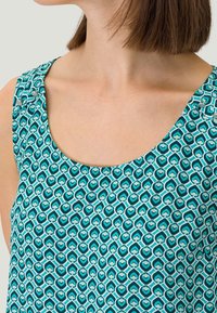 Grønblåt tanktop med et geometrisk mønster af hjerter med hvide konturer. Den runde halsudskæring og knapperne tilføjer detaljer til skulderen.