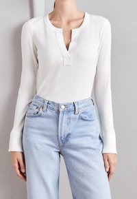 Vit ribbad långärmad topp med henley-krage och en liten knappdetalj, i kombination med ljusblå högmidjade denimjeans.