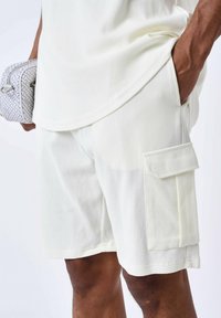 Pantalones cortos color crema hechos de una tela texturizada con un bolsillo cargo en el lateral, combinados con una parte superior a juego. Diseño minimalista sin patrones.