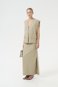Gilet di lino beige con spalle ampie e scollo a V, abbinato a una gonna midi coordinata con uno spacco laterale. Piccola borsa a tracolla nera con motivo.