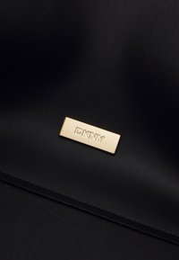 Svart lærveske med en gull rektangulær tag gravert med "DKNY," som fremhever dens teksturerte overflate og elegante design.