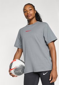 Szara krótka koszulka z krótkim rękawem z czerwonym logo Nike, zestawiona z czarnymi legginsami, trzymająca wzorzystą piłkę nożną w czerwono-czarnym kolorze.