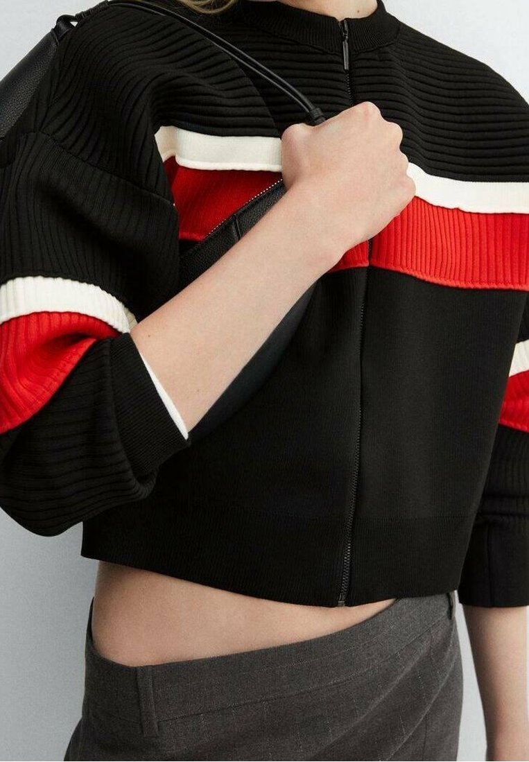 Pull court noir côtelé avec des accents rayés rouges et blancs, col rond et fermeture éclair à l'avant. Porté avec un pantalon gris.