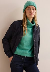 Marineblauer Bomberjacke mit Druckknöpfen über einem türkisfarbenen Rollkragenpullover. Das Modell trägt zudem eine gerippte türkisfarbene Mütze. Dunkle Jeans vervollständigen das Outfit.