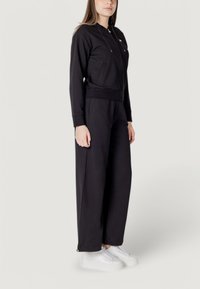 Tuta nero in tessuto morbido con felpa con zip, vita elasticizzata e pantaloni a gamba larga. Abbinato a sneakers bianche con platform.