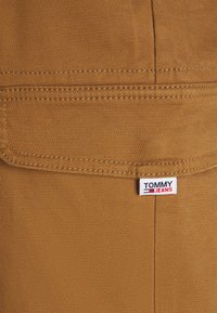 Tissu marron avec une texture lisse, comportant une poche cousue et une étiquette "TOMMY JEANS" visible aux couleurs rouge, blanc et bleu.