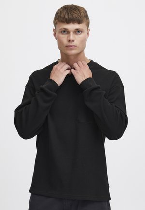 Camisa de manga larga negra hecha de tejido de punto waffle texturizado, con un escote redondo y un bolsillo en el pecho. Ajuste casual y relajado.