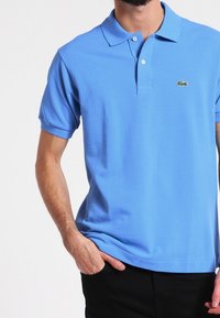 Ljust blå pikétröja av mjukt tyg, med klassisk krage, två knappar och en liten grön Lacoste-logga på vänster bröst.