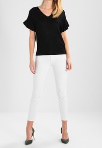 Žena oblečená v černém krátkém rukávu V-krku, bílých slim-fit kalhotách a tmavě zelených lodičkách stojící na bílém pozadí.