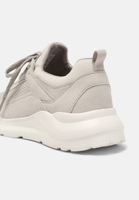 Baskets gris clair avec une tige en suède texturé et tissu, une semelle blanche épaisse et une languette à l'arrière. Des lacets sont visibles à l'avant.