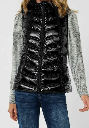Frau trägt eine glänzende schwarze gesteppte Weste über einem grauen gestreiften Langarmshirt und blauen Jeans, gezeigt vom Hals bis zu den Hüften.