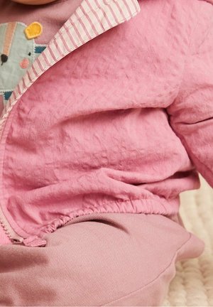 Niemowlę ubrane w różową pikowaną kurtkę z paskowanymi lamówkami oraz koszulę z kolorową naszywką z zwierzęciem, siedzące na miękkiej, teksturowanej powierzchni.