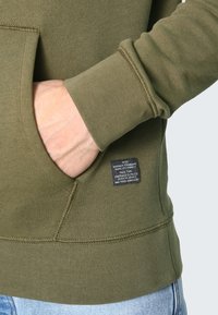 Felpa verde oliva con tasca a marsupio, polsini a costine e etichetta in tessuto sulla cucitura laterale. Tessuto morbido e strutturato con cuciture visibili.