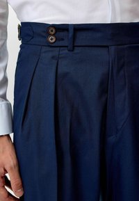 Marineblaue Hosen mit Fischgrätenstruktur, ausgestattet mit einem doppelten Knopfverschluss, Gürtelschlaufen und scharfen Vorderfalten.