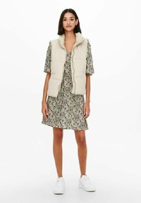 Gilet rembourré beige avec col haut sur une courte robe à motifs avec un design paisley. Le mannequin porte des baskets blanches.