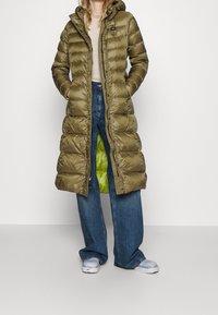 Abrigo largo acolchado de color verde oliva con capucha, botones a presión y bolsillos con cremallera. El interior tiene un forro verde brillante. Llevado sobre vaqueros azules.