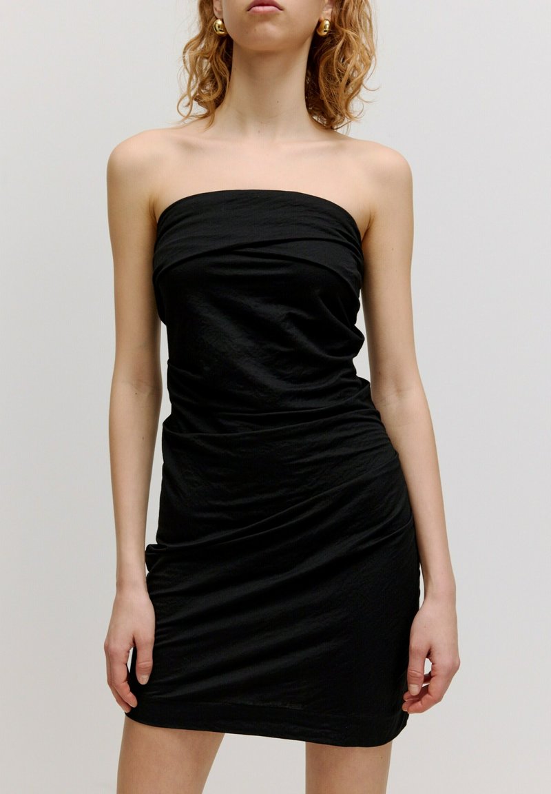 Zwarte strapless jurk van kreukelig materiaal. Aansluitend ontwerp met geplooide details bij de taille, tot halverwege de dij. Glad textuur.