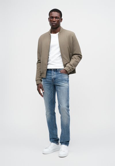 Ruda bomberio striukė, balta marškinėliai, šviesiai mėlyni džinsai ir balti sportbačiai. Lygi striukės tekstūra, įprastos dydžio fasonas, standartinė džinsų kirpimo forma.