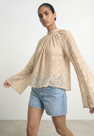 Kvinde iført en beige bluse med lange ærmer i blonder med vide ærmer og lyseblå denimshorts mod en ensfarvet baggrund.