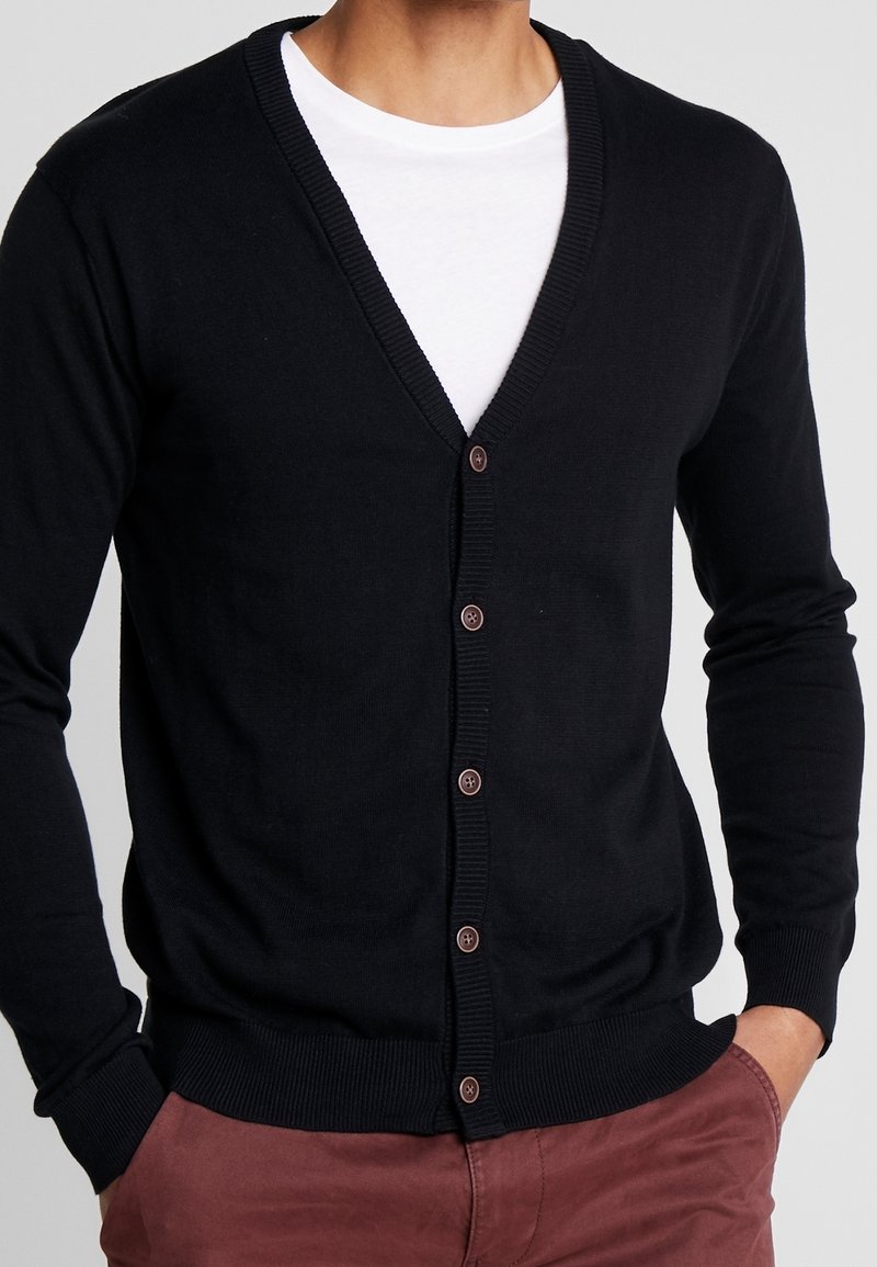 Homme portant un cardigan noir boutonné sur un t-shirt blanc, les mains dans les poches, assorti à un pantalon bordeaux.