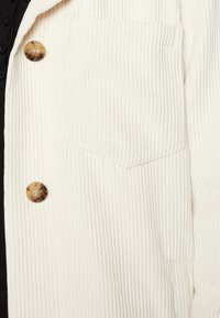 Giacca in corduroy bianco con righe verticali, dotata di una tasca sul petto e di un bottone rotondo in colore tartaruga. Texture liscia e design casual.
