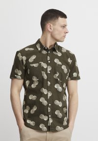 Camisa de manga corta de color oliva con un patrón de conchas crema, cierre de botones en el frente, dos bolsillos en el pecho y un dobladillo redondeado. Confeccionada en una tela ligera.