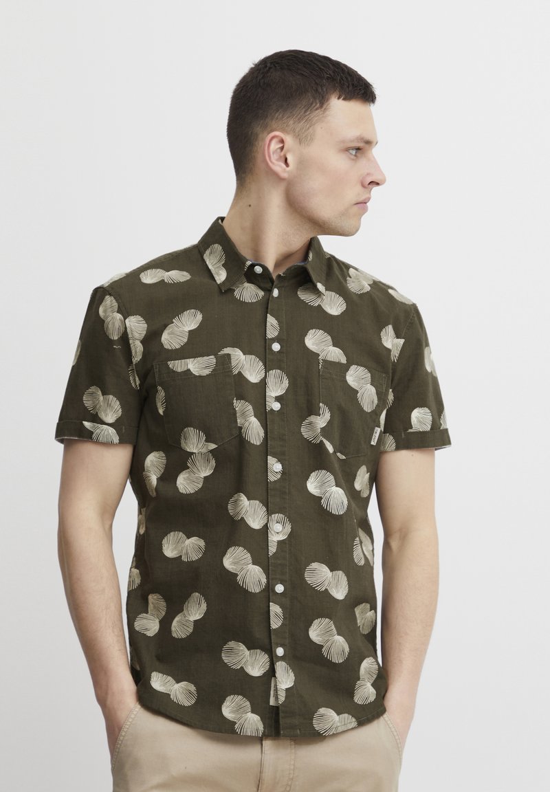 Camisa de manga corta de color oliva con un patrón de conchas crema, cierre de botones en el frente, dos bolsillos en el pecho y un dobladillo redondeado. Confeccionada en una tela ligera.
