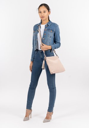 Light pink handbag made of textured material, featuring a long strap and tassel detail. Worn by a person in denim jacket and jeans.