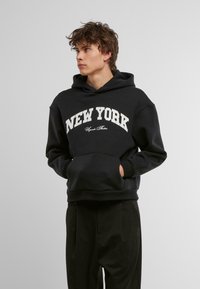 Zwarte hoodie van zacht materiaal met een kangaroo-pocket en witte tekst "NEW YORK" in een gebogen ontwerp. Casuale pasvorm met een capuchon.