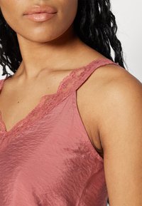 Hihaton vaaleanpunainen toppi, jossa on pehmeä, hieman ryppyinen pinta, ja joka sisältää pitsireunuksen kaulanauhassa ja olkaimissa lisäyksinä.