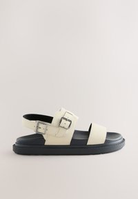 Sandale en cuir beige avec deux boucles argentées réglables, bout et talon ouverts, et une semelle noire épaisse.