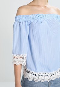 Blouse bleu clair à rayures, décolletée, avec manches courtes bouffantes et bordure en dentelle blanche à l'ourlet. Tissu doux et léger ; design décontracté.