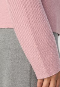 Gros plan sur la main d'une personne reposant le long du corps, portant un pull en maille rose texturé et un pantalon en laine gris.