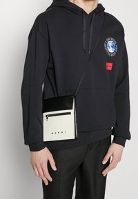 Sweat à capuche noir avec impression graphique et logo rouge, porté avec un sac bandoulière en cuir noir et blanc avec fermeture éclair et marquage.