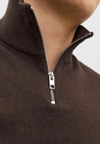 Pull en tricot brun avec un col montant et une fermeture éclair argentée ; texture lisse ; gros plan sur la zone du cou mettant en avant le détail de la fermeture éclair.
