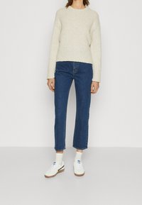 Maglione beige lavorato a maglia con maniche larghe, abbinato a jeans a gamba dritta blu medio e sneaker bianche con dettagli marroni.