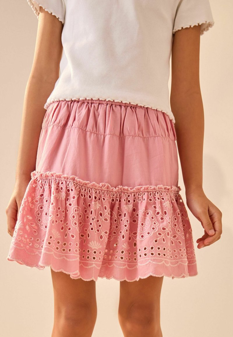 Jupe en coton rose avec un ourlet à volants, ornée d'une broderie délicate en œillets et d'une taille froncée. Associée à un dessus blanc côtelé.