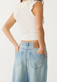 Vit stickad ärmlös topp med vågig krage, i kombination med ljusblå vida denimjeans med två bakfickor och en läderetikett.