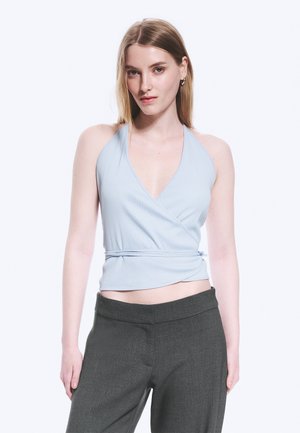 Νεαρή γυναίκα με ανοιχτό μπλε αμάνικο wrap τοπ και σκούρο γκρι παντελόνι στέκεται μπροστά σε απλό λευκό φόντο.