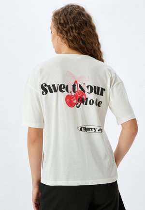 T-shirt en coton blanc avec un grand motif de cerises rouges accompagné d'un ruban rose, et le texte "Sweet Sour Mode" en lettres noires en gras.
