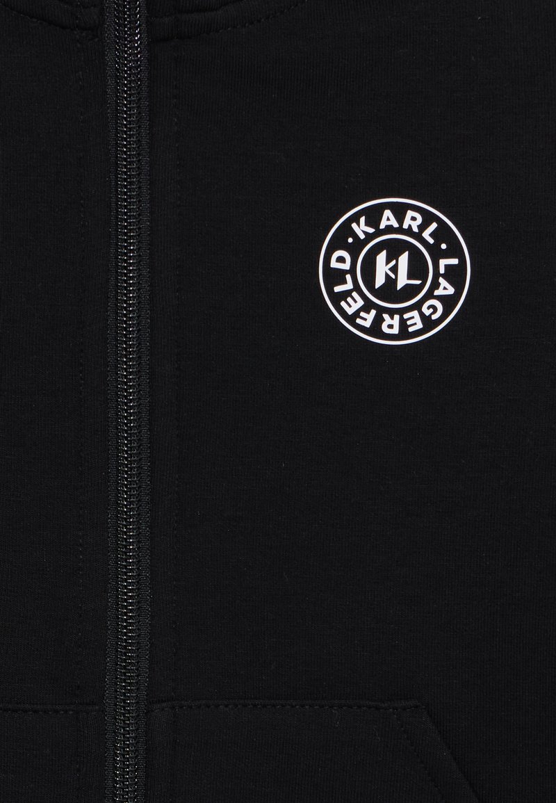 Haut à zip noir en tissu doux avec une texture côtelée. Présente un logo blanc circulaire avec "Karl Lagerfeld" et les initiales "KL."
