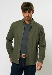 Olīvu zaļš pogājams krekls ar izplestu apkakli, gariem piedurknēm un netraucētu logotipu. Nēsāts virs strukturēta pelēka T-kreklu, kombinēts ar tumšām džinsām.