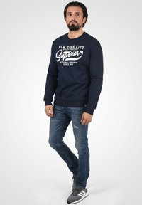 Mørkeblå sweatshirt med hvid tekst, lange ærmer, ribbede manchetter og kant. Sammen med slidte blå jeans og grå sportive sko.