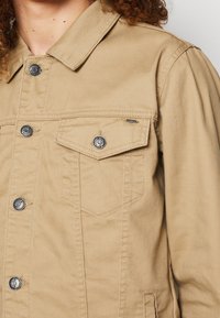 Beżowa kurtka dżinsowa z kołnierzykiem, zatrzaskami i kieszenią na piersi. Charakteryzuje się strukturalnym fasonem i teksturowanym materiałem.