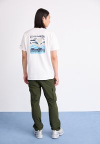 Camiseta blanca con un gráfico colorido de una escena montañosa en la parte posterior, combinada con pantalones cargo verdes y zapatillas de deporte gris claro.