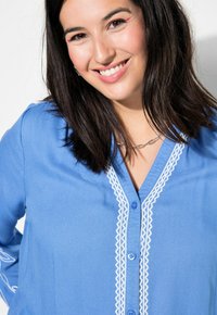 Camicia azzurra con bottoni realizzata in tessuto liscio, con dettagli ricamati bianchi lungo la patta e i polsini, e design a scollo a V.