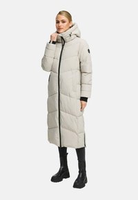Donna che indossa un lungo cappotto imbottito beige chiaro con cappuccio, cerniera nera, stivali neri e pantaloni scuri, in piedi contro uno sfondo neutro.