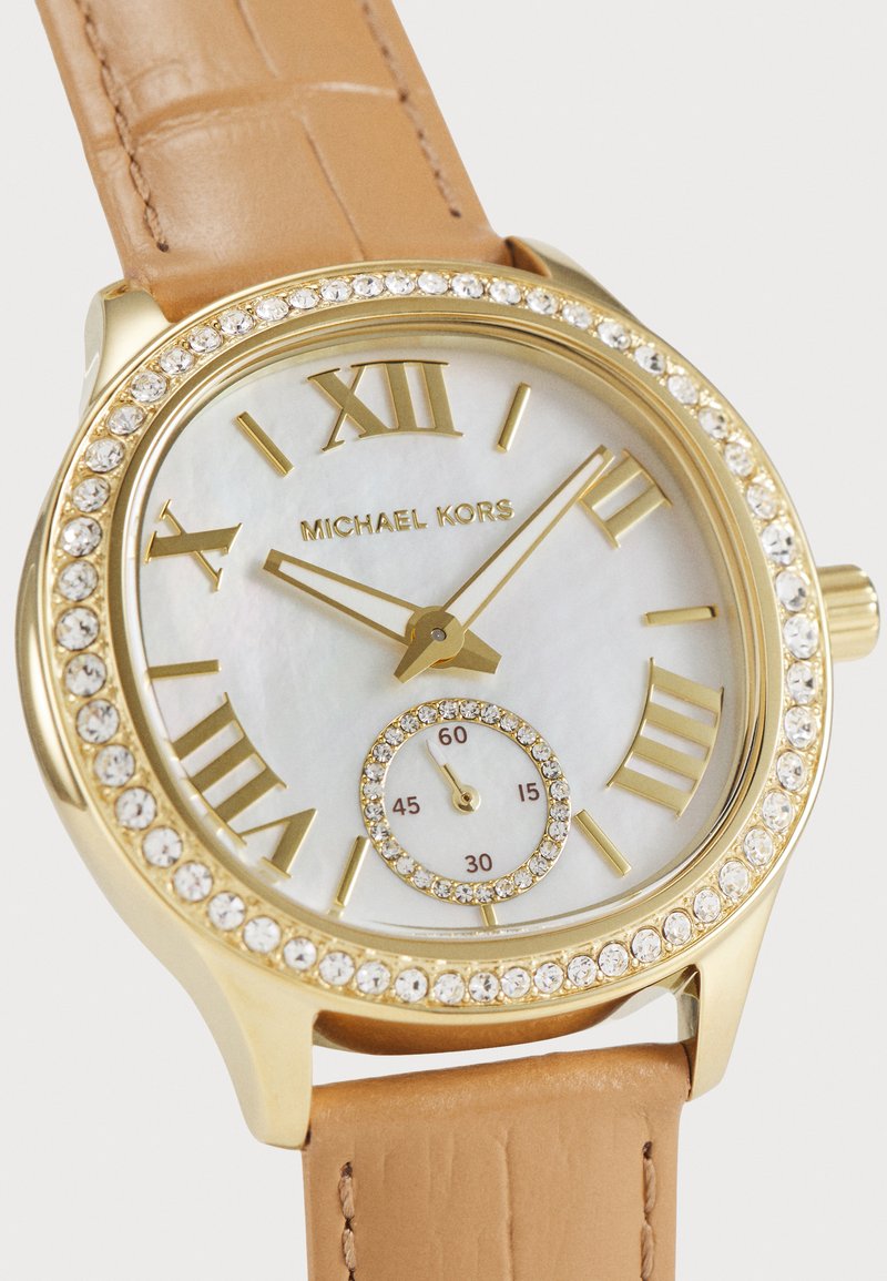 Montre dorée avec un cadran en nacre, chiffres romains et un boîtier serti de cristaux. Équipée d'un bracelet en cuir beige et d'un sous-cadran.